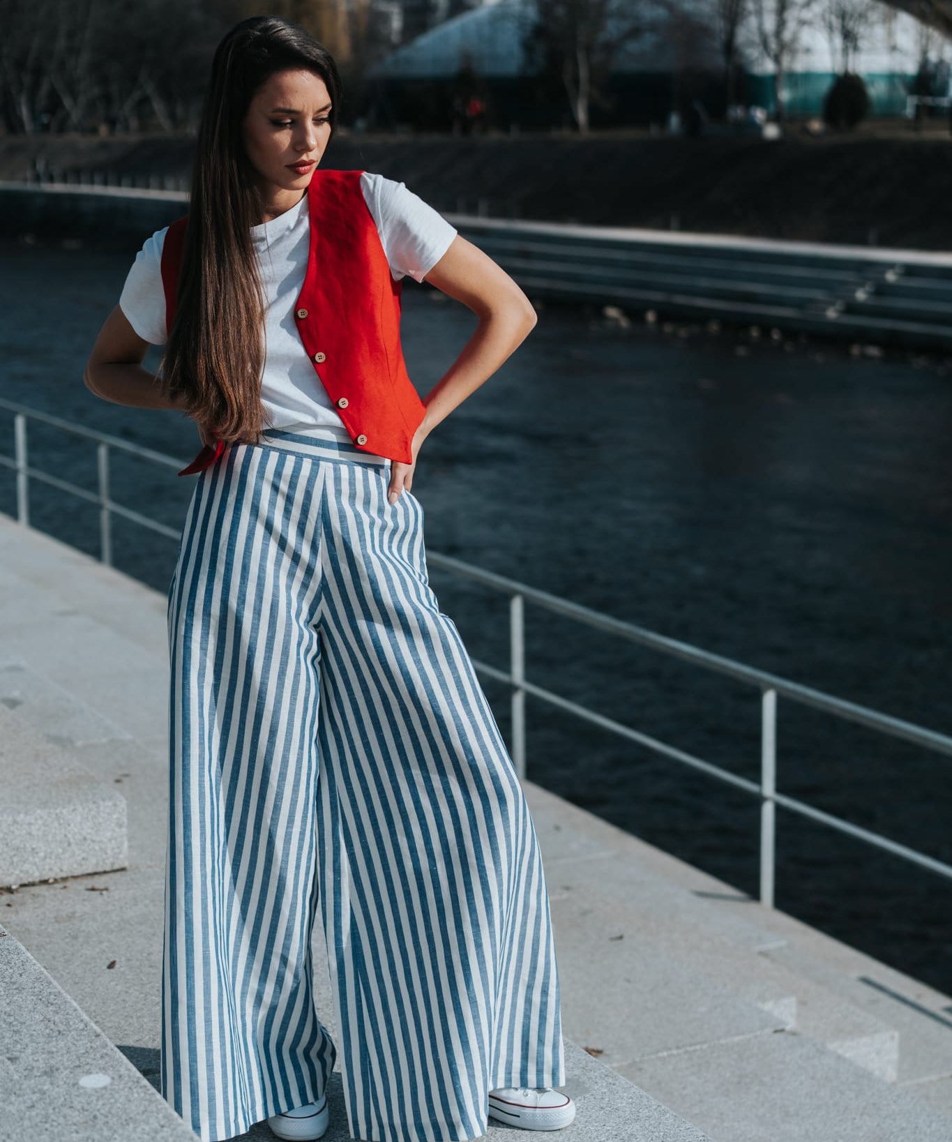 Woman wearing a white linen top, red linen vest, and striped wide leg linen pants by a waterfront.